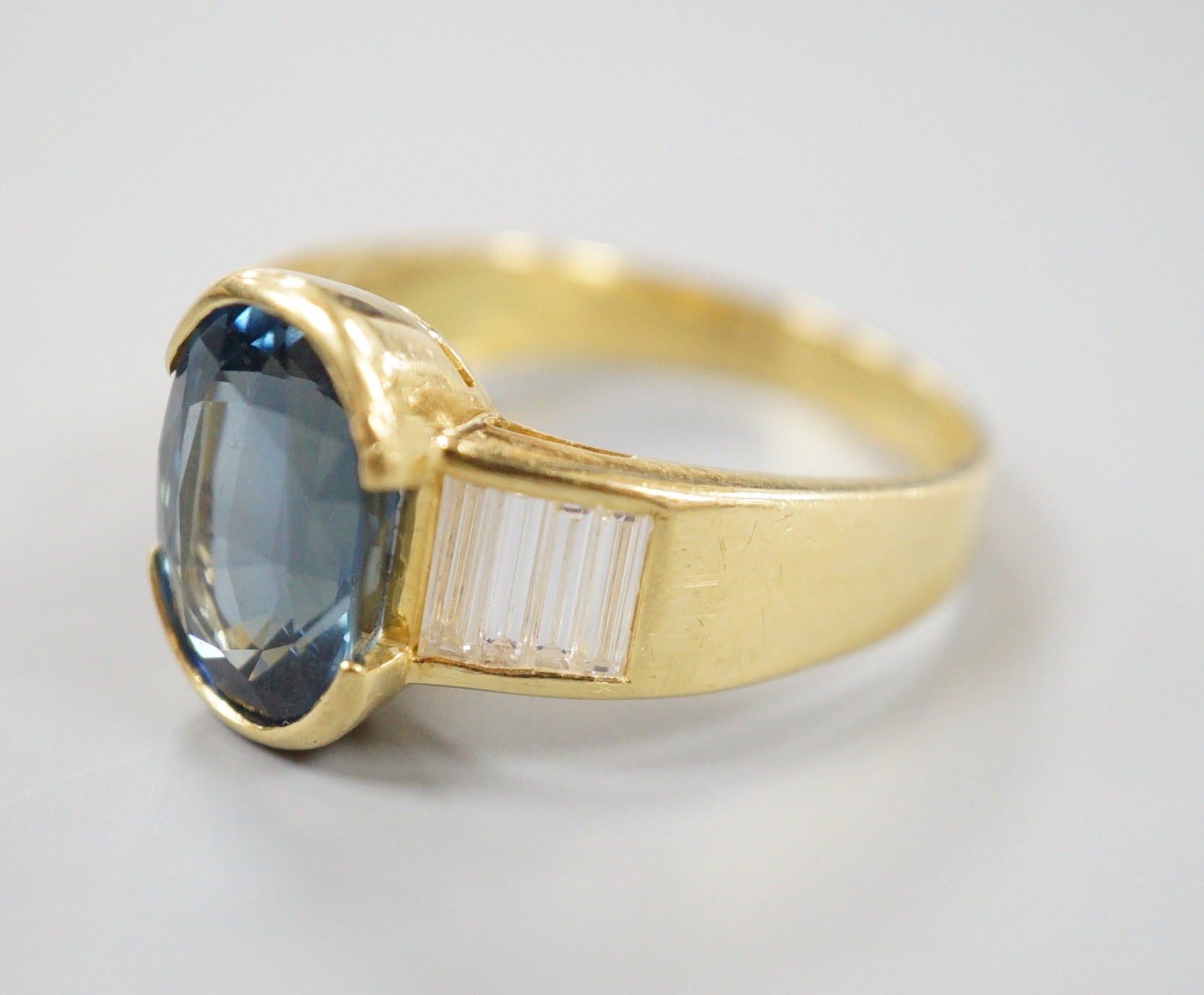 A modern 18ct gold and single stone oval cut sapphire set ring, with baguette cut diamond set shoulders, size R, gross weight 8.3 grams.